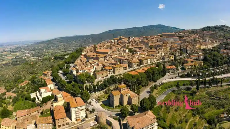 A long wedding reception table set under fairy lights in a Tuscan vineyard at sunset, with Malaysian and Italian guests mingling.