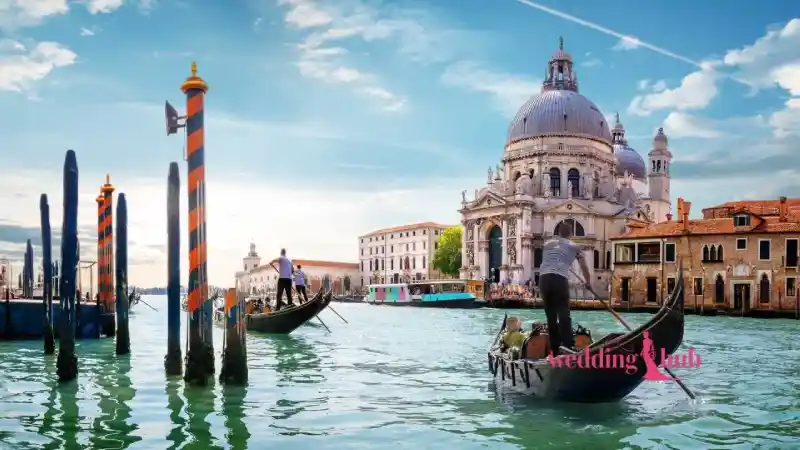 A Malaysian bride and groom in traditional and western attire smiling in a gondola on a canal in Venice, Italy, with historic buildings in the background.